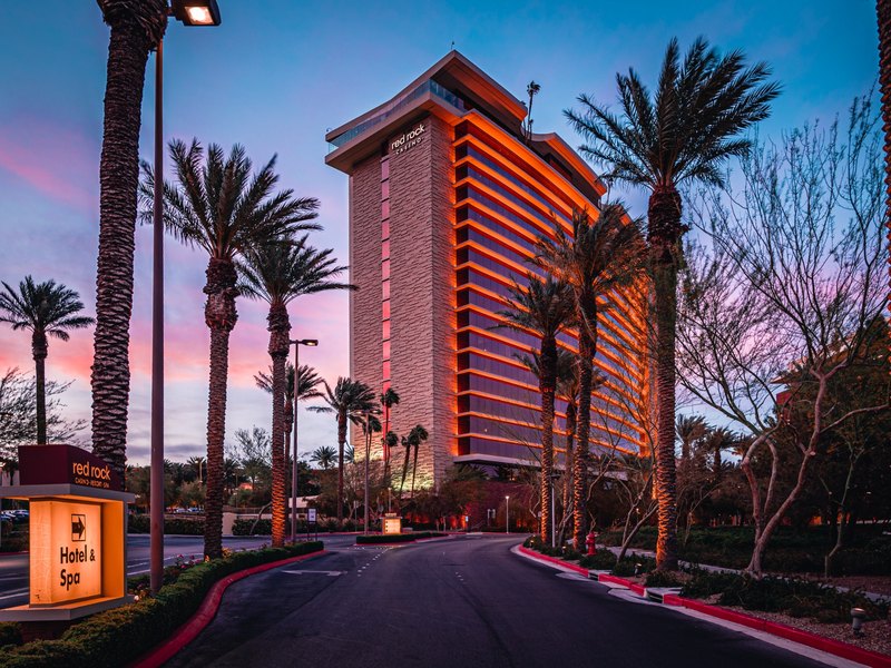 Red Rock Casino Resort & Spa rises against a colorful desert sunset, framed by palms along the hotel entrance drive.