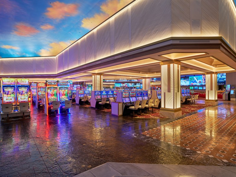 A bright, spacious section of the Sunset Station casino floor features rows of slot machines beneath modern illuminated architectural details.