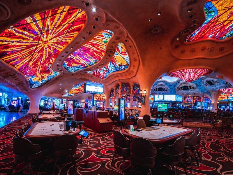 Table games sit beneath vivid stained-glass-style ceiling panels, showcasing Sunset Station’s colorful casino design and energetic gaming floor.